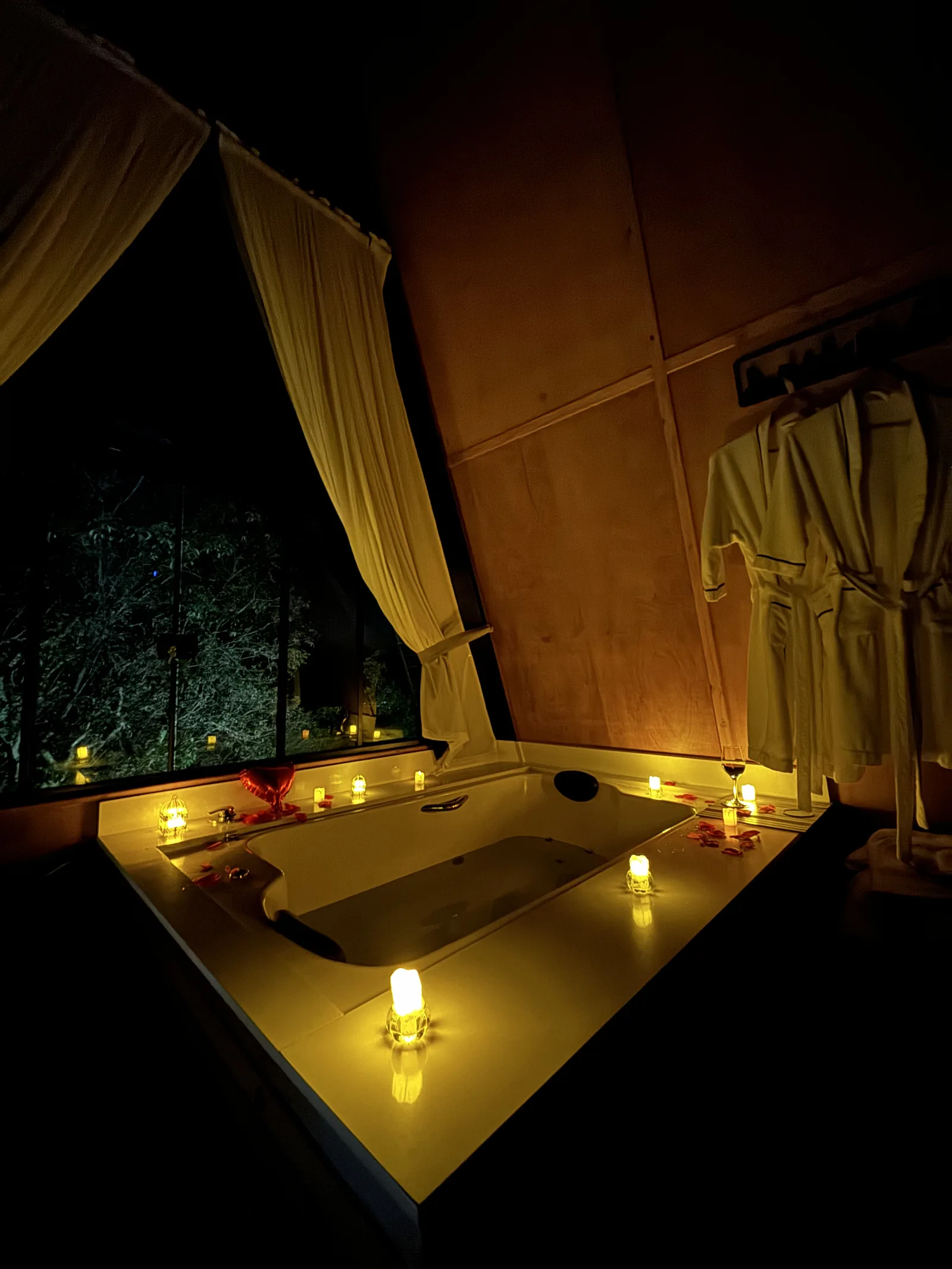 Banheira decorada com velas e pétalas à noite, vista para o mato